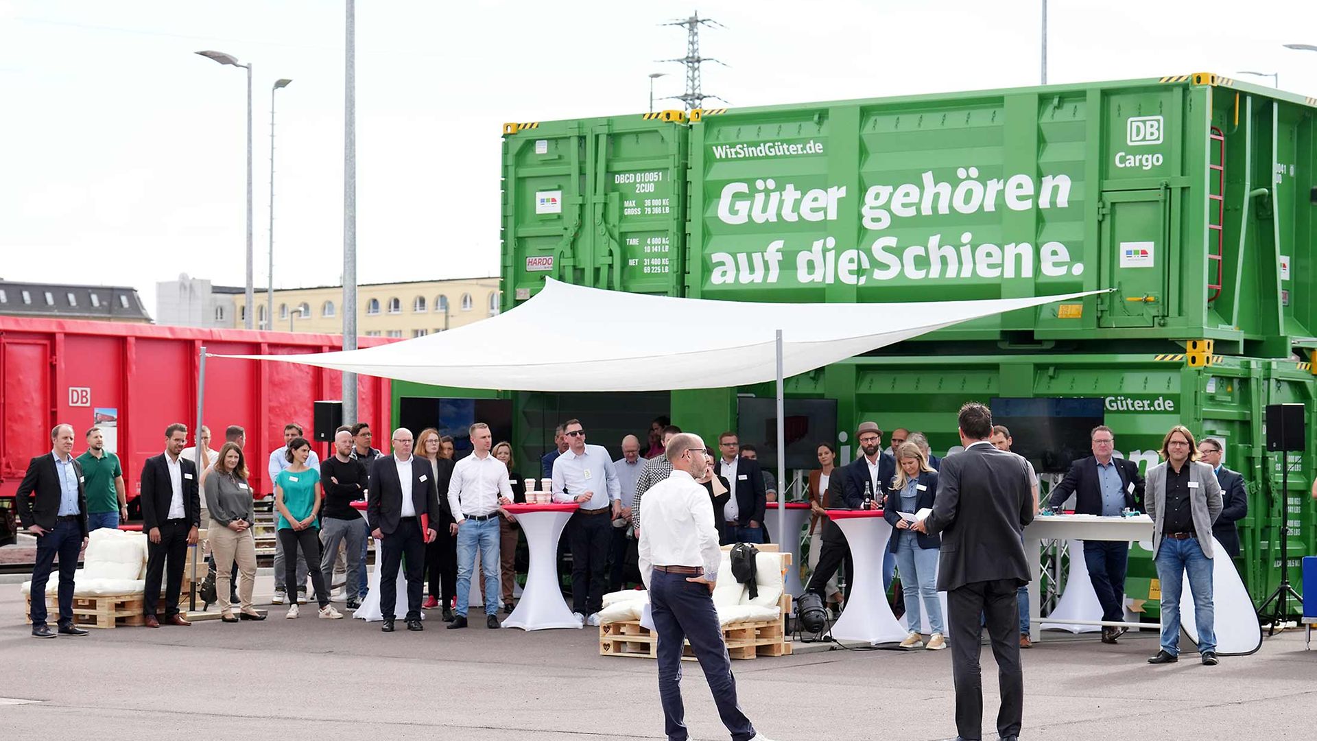 Vor 2 gestapelten grünen Containern stehen Teilnehmer eines Termins an Stehtischen.