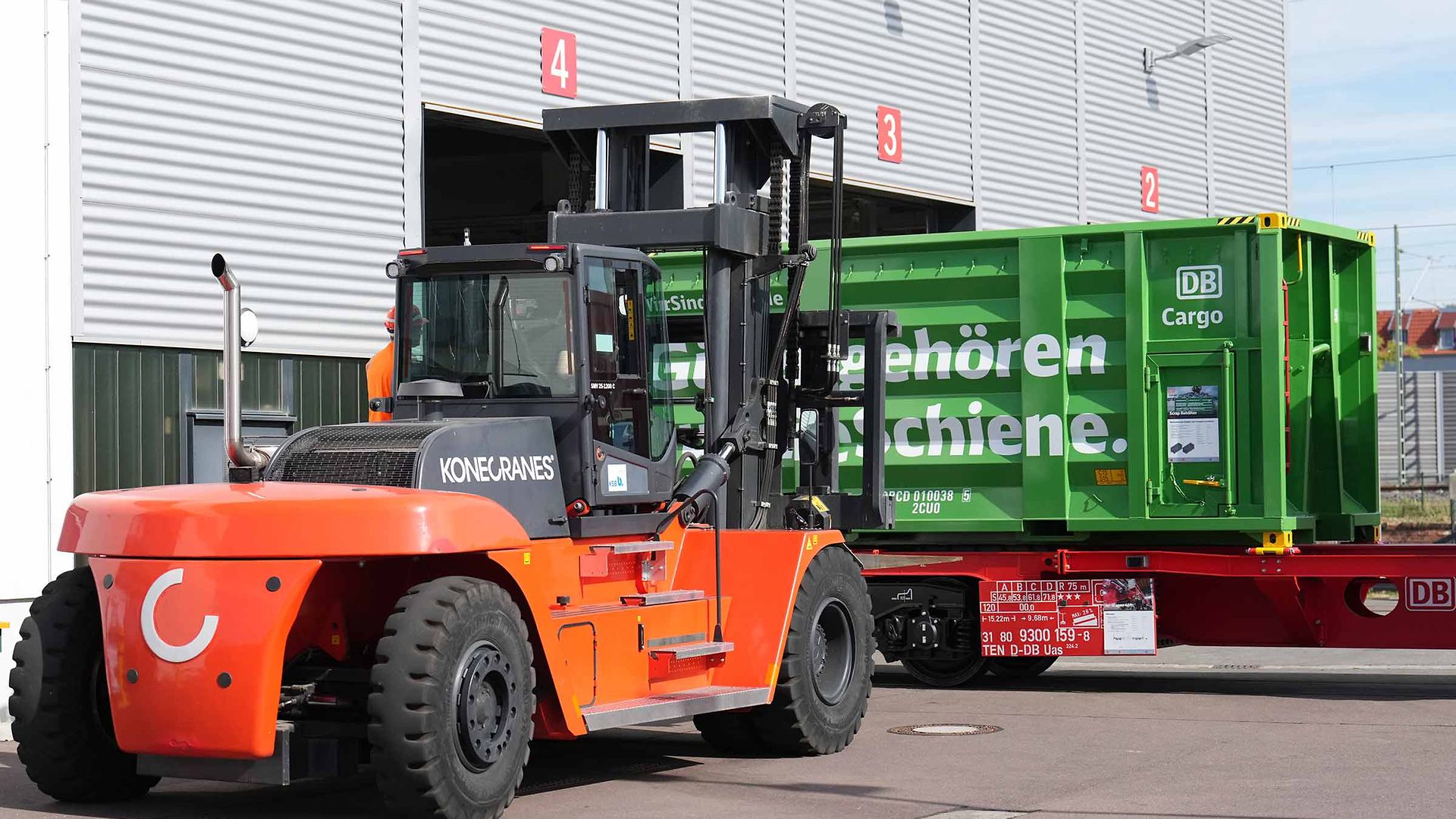 A green container is transported by a forklift truck and loaded onto a freight wagon.