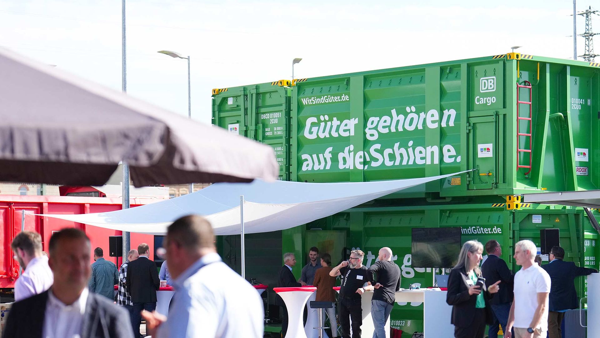 Discussion groups of an appointment stand under sunshades in front of 2 stacked green containers.