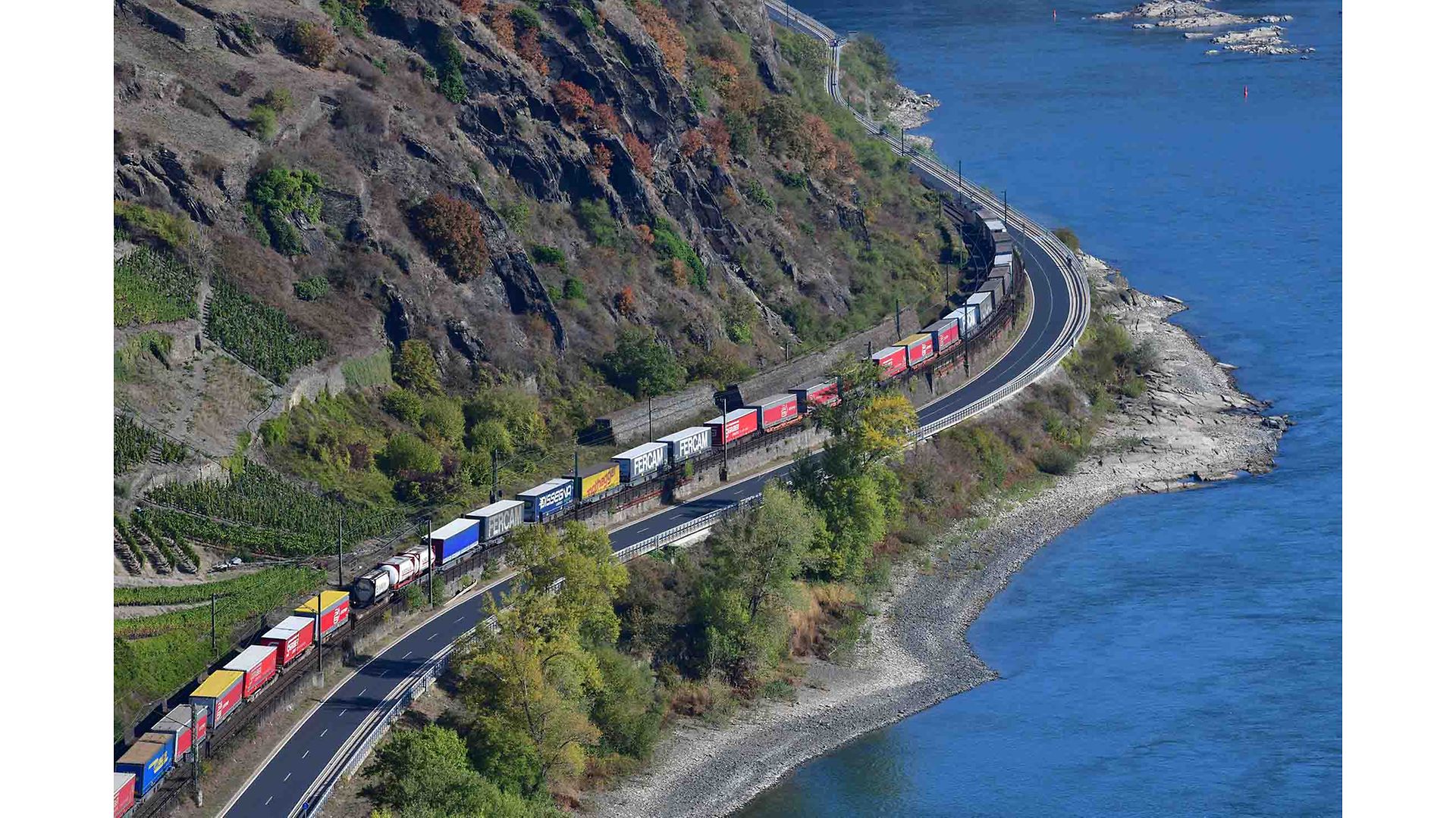 Ein Zug mit verschiedenen Containern fährt an einem Fluss entlang.