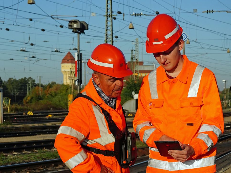 Zwei Männer in orangefarbener Schutzkleidung besprechen sich im Gleisbett an einem Rangierbahnhof.