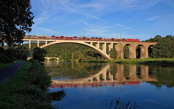 Ein Güterzug mit mehreren Güterwagen fährt unter blauem Himmel über eine Brücke.