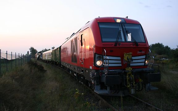 Rote Zweikraftlok fährt mit Güterwagen in Richtung Kamera unter blauem Himmel.