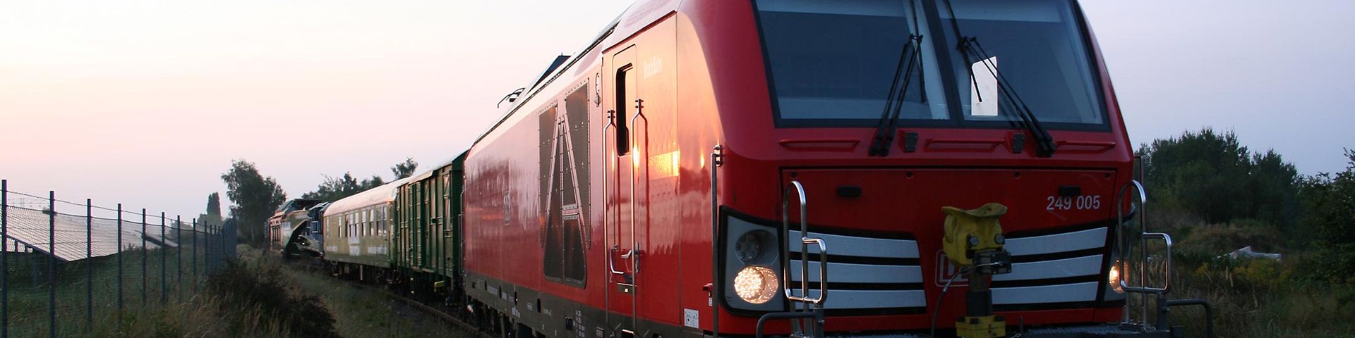 Rote Zweikraftlok fährt mit Güterwagen in Richtung Kamera unter blauem Himmel.