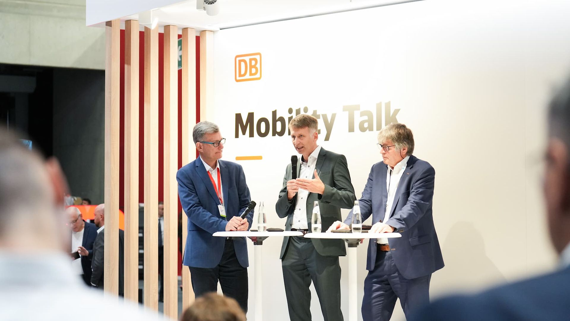 Drei Personen stehen mit Mikrophonen auf einer Bühne bei einer Podiumsdiskussion. Im Hintergrund steht „DB Mobility Talk“.