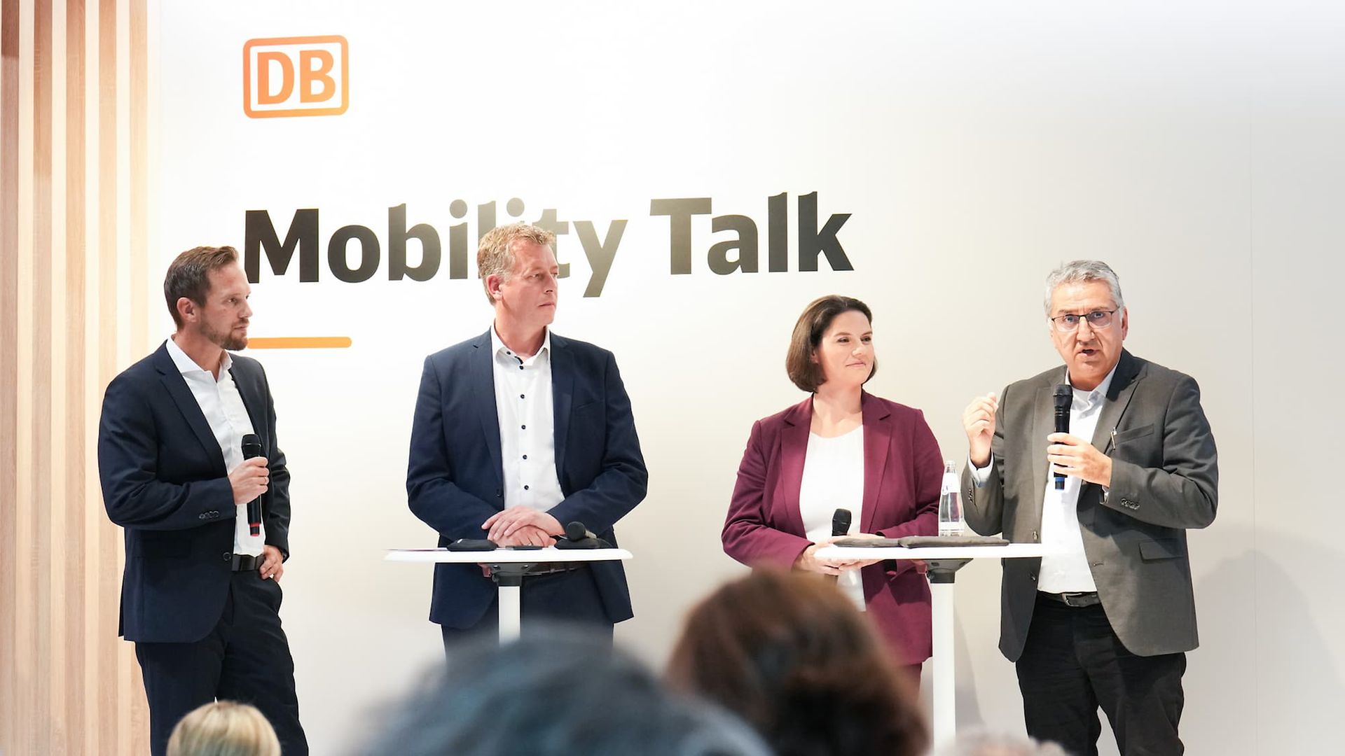 Vier Personen stehen mit Mikrophonen auf einer Bühne bei einer Podiumsdiskussion. Im Hintergrund steht „DB Mobility Talk“.