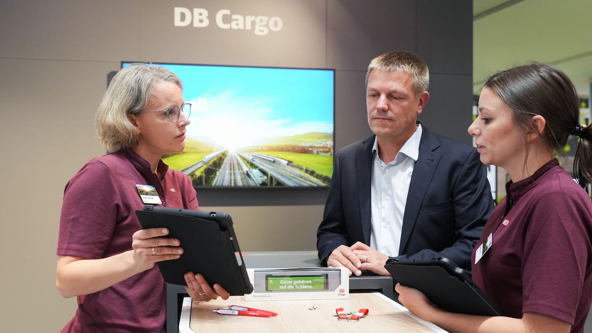 Three people stand with a tablet at the DB Cargo stand at InnoTrans 2024.