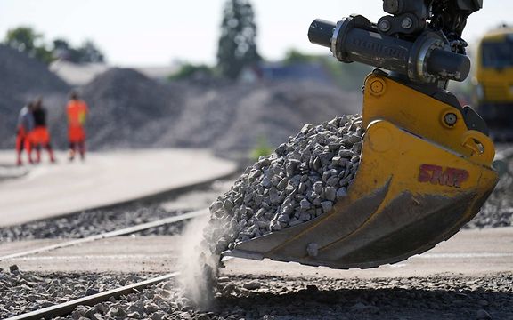 Bagger schüttet Schotter auf Gleise, im Hintergrund Arbeiter in Warnkleidung.