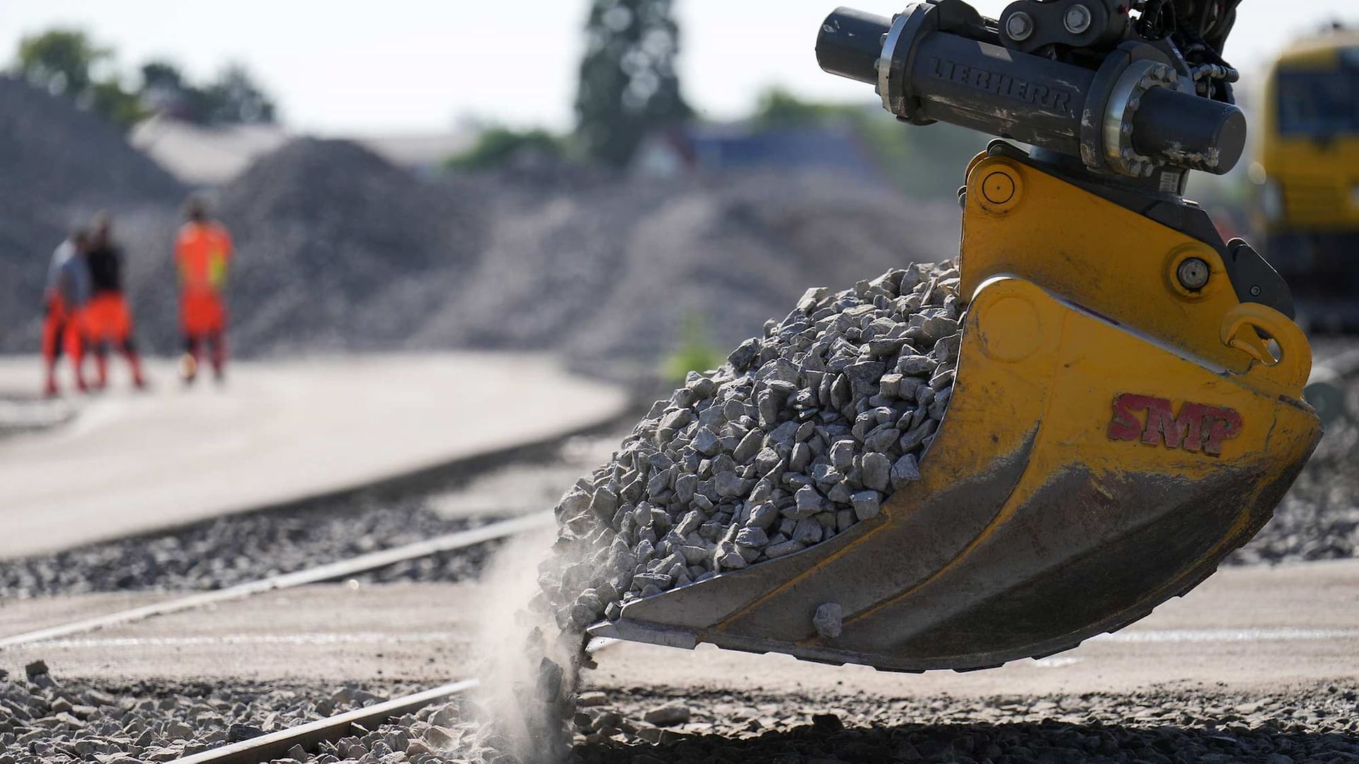 Bagger schüttet Schotter auf Gleise, im Hintergrund Arbeiter in Warnkleidung.