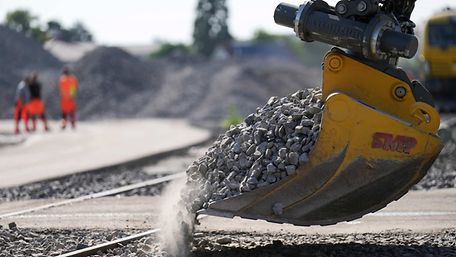 Excavator pours ballast onto tracks, in the background workers in high-visibility clothing.