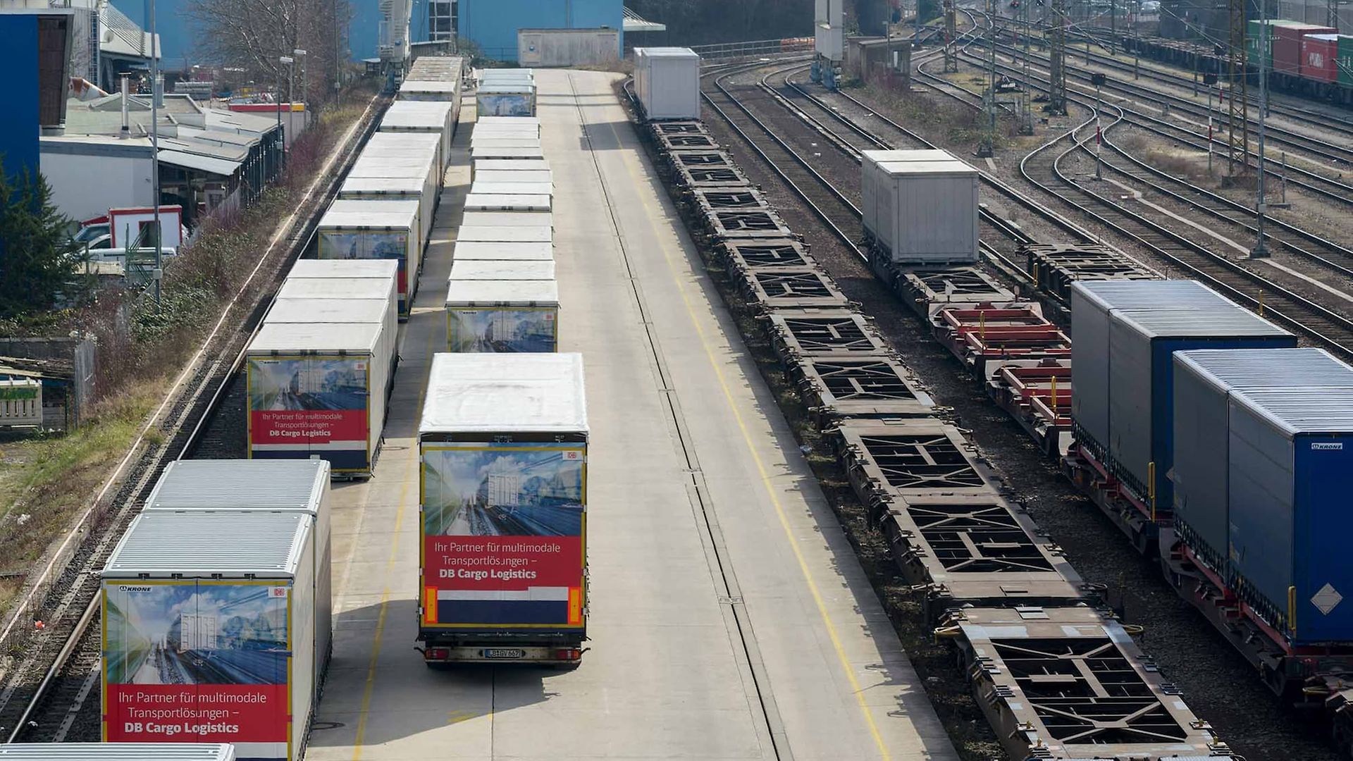 Lkw mit Containern, leere Containertragwagen und Tragwagen mit Containern stehen in einem Umschlagbahnhof nebeineinander.