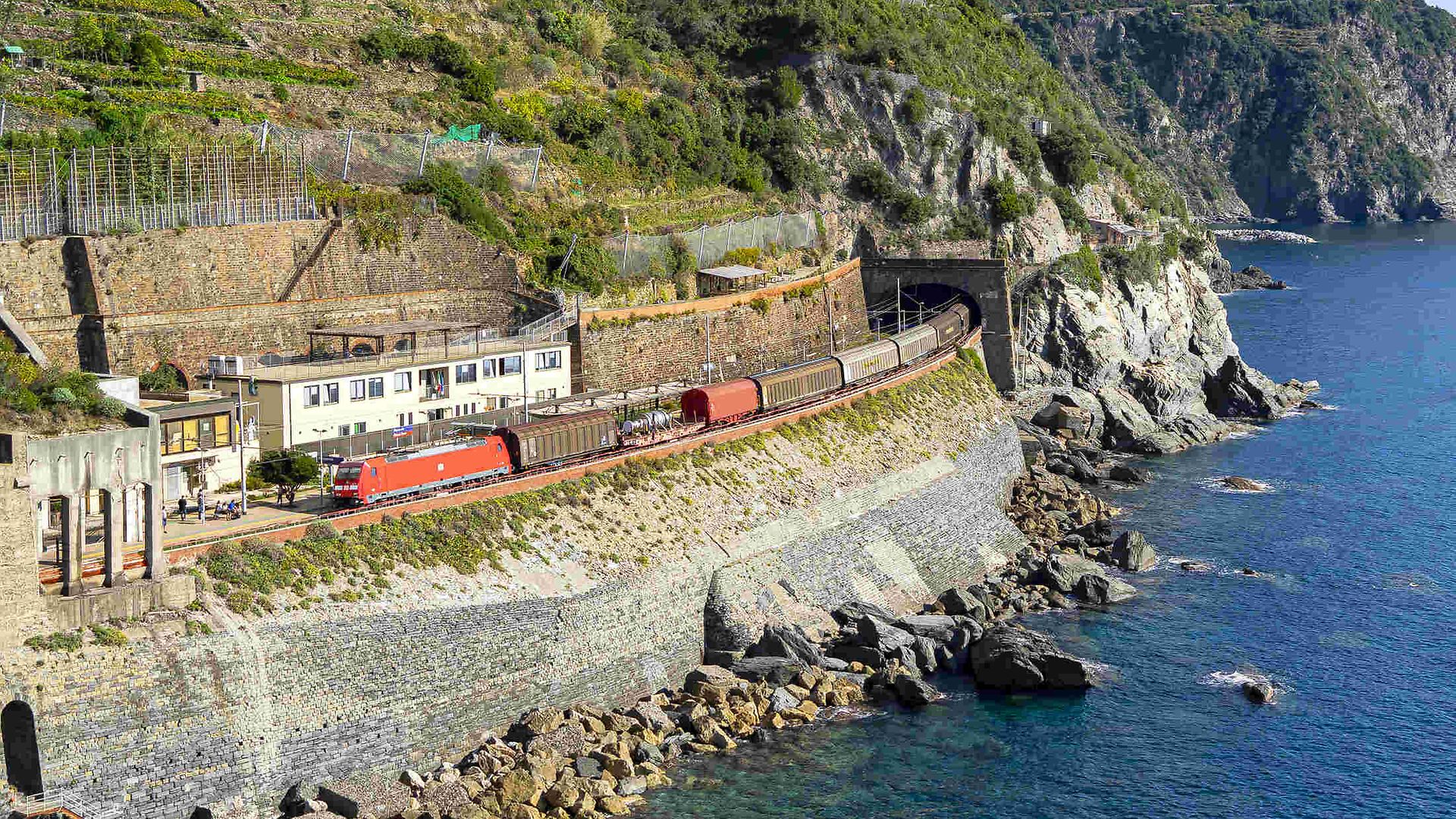 Freight train travelling along the Italian coast and just coming out of a tunnel, with the sea on the right.