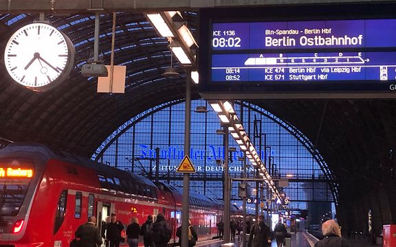 Bahnhof am Morgen mit Uhr, Anzeigetafel, rotem Zug und Passanten.