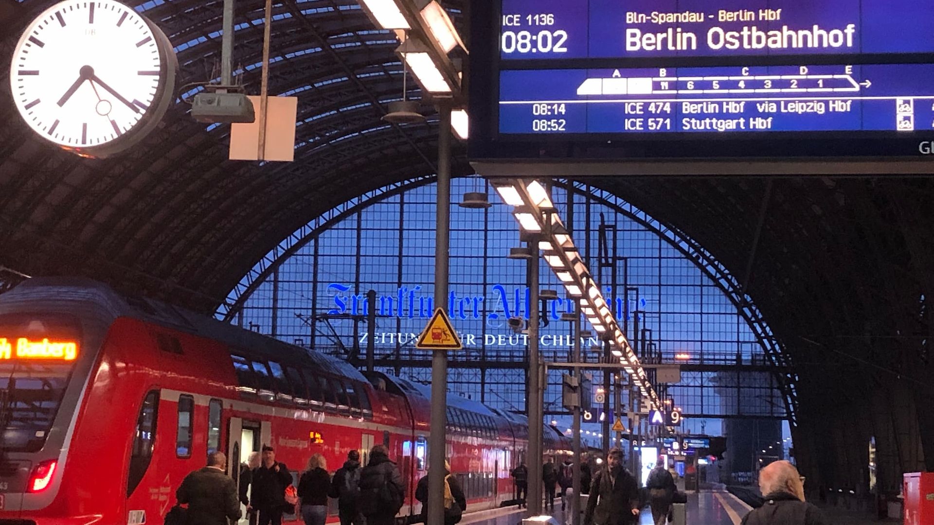 Bahnhof am Morgen mit Uhr, Anzeigetafel, rotem Zug und Passanten.