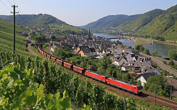 Güterzug mit roten Loks fährt durch Weinberge und ein Dorf an der Mosel, mit Fluss und Hügeln im Hintergrund.