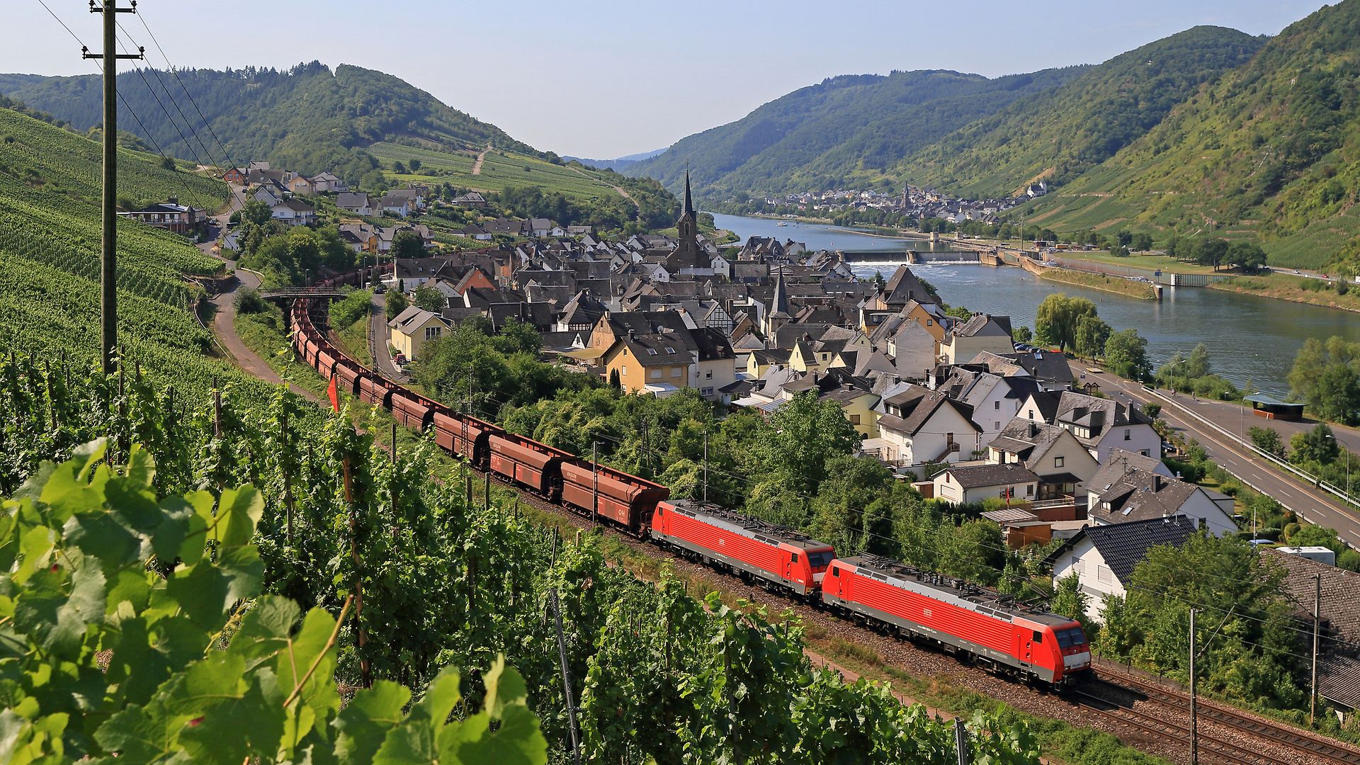 Freight train with red locomotives travelling through vineyards and a village on the Moselle, with river and hills. 