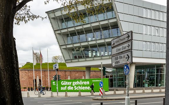 Moderne DB-Cargo-Zentrale in Mainz mit grünem Werbecontainer und Schildern zu lokalen Zielen.