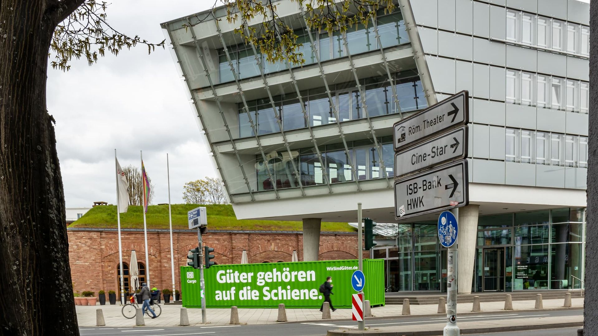 Moderne DB-Cargo-Zentrale in Mainz mit grünem Werbecontainer und Schildern zu lokalen Zielen.