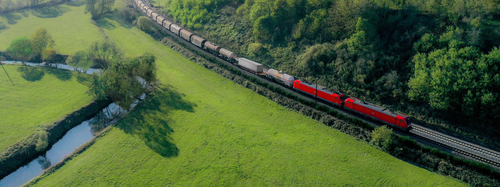 Roter DB Cargo Güterzug fährt durch eine grüne Hügellandschaft mit Wäldern, Wiesen und einem kleinen Bach