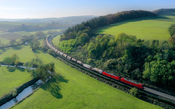 Red DB Cargo freight train travelling through a green hilly landscape with forests, meadows and a small stream