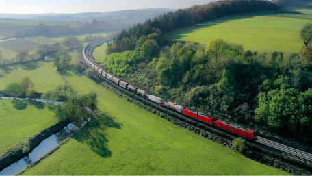 Red DB Cargo freight train travelling through a green hilly landscape with forests, meadows and a small stream