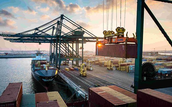 Harbour with cranes, container ship at the quay, crane lifting containers. Stacked containers and vehicles in the background.