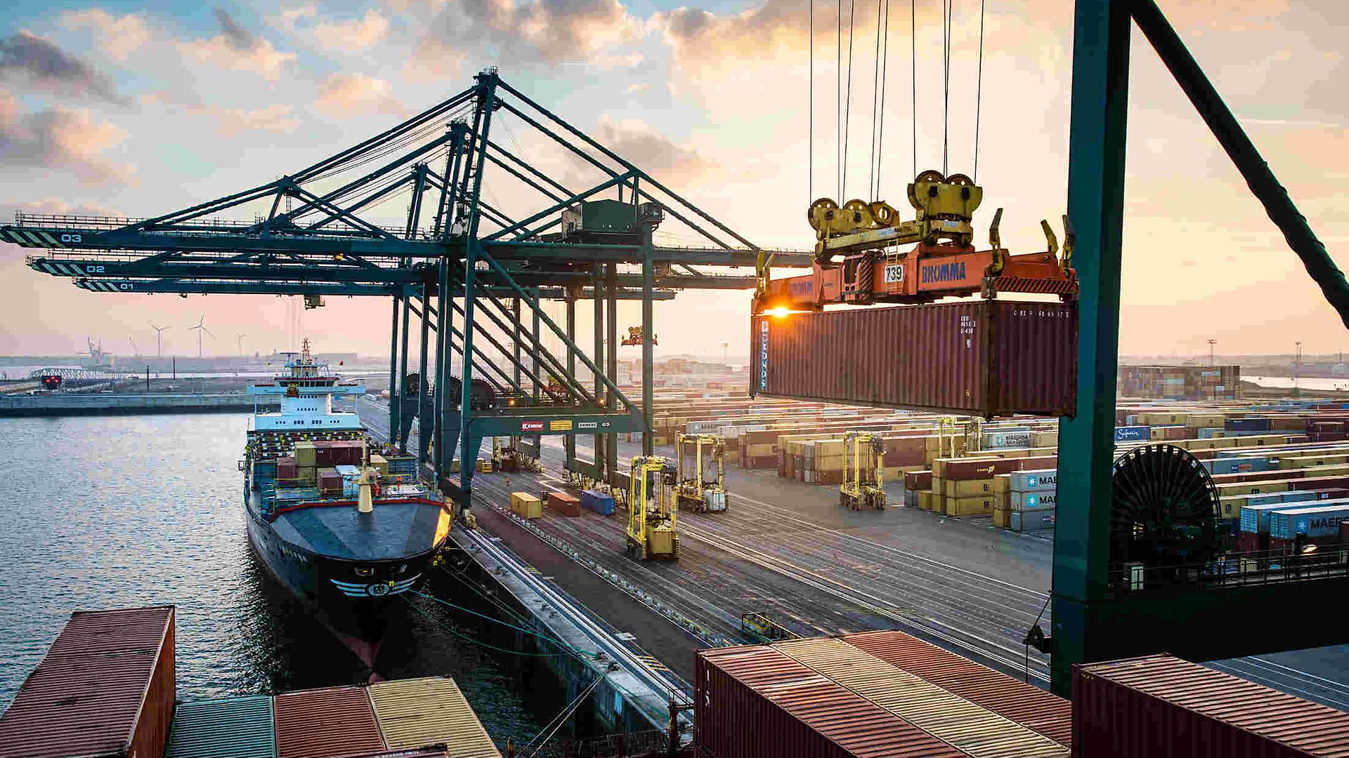 Harbour with cranes, container ship at the quay, crane lifting containers. Stacked containers and vehicles in the background.