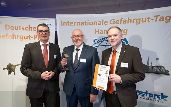 Drei Männer in Anzügen bei der Verleihung des Deutschen Gefahrgut-Preises 2025, einer hält die Trophäe, ein anderer die Urkunde.