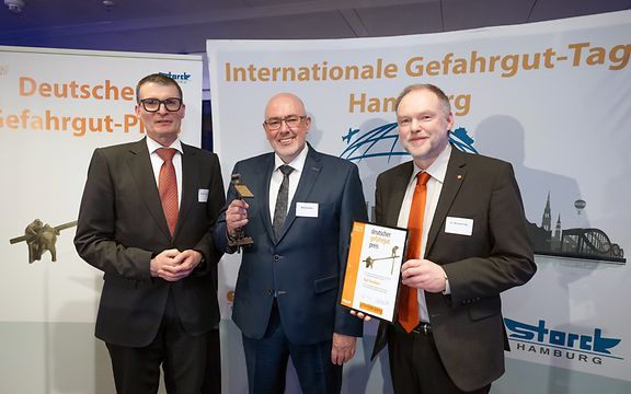 Three men in suits at the German Dangerous Goods Award 2025 ceremony, one holding the trophy, another the certificate.