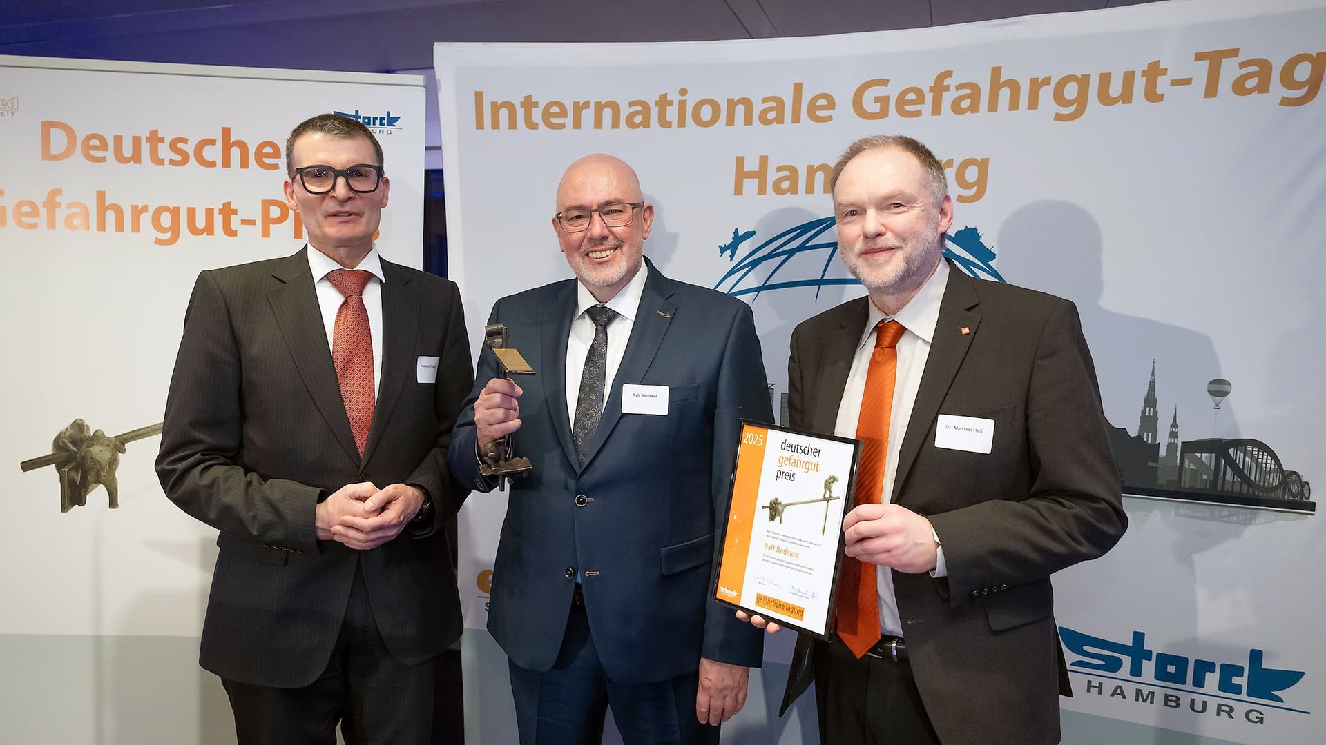 Three men in suits at the German Dangerous Goods Award 2025 ceremony, one holding the trophy, another the certificate.